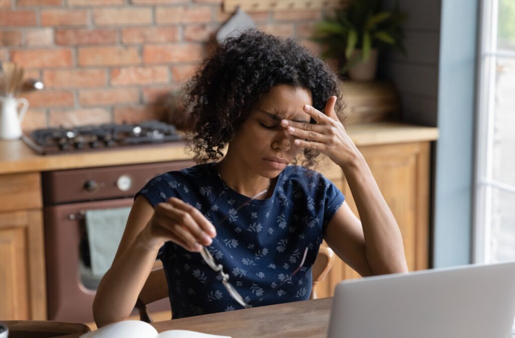 A woman with dark curly hair in front of a laptop, taking off her glasses and rubbing the space between her eyes.