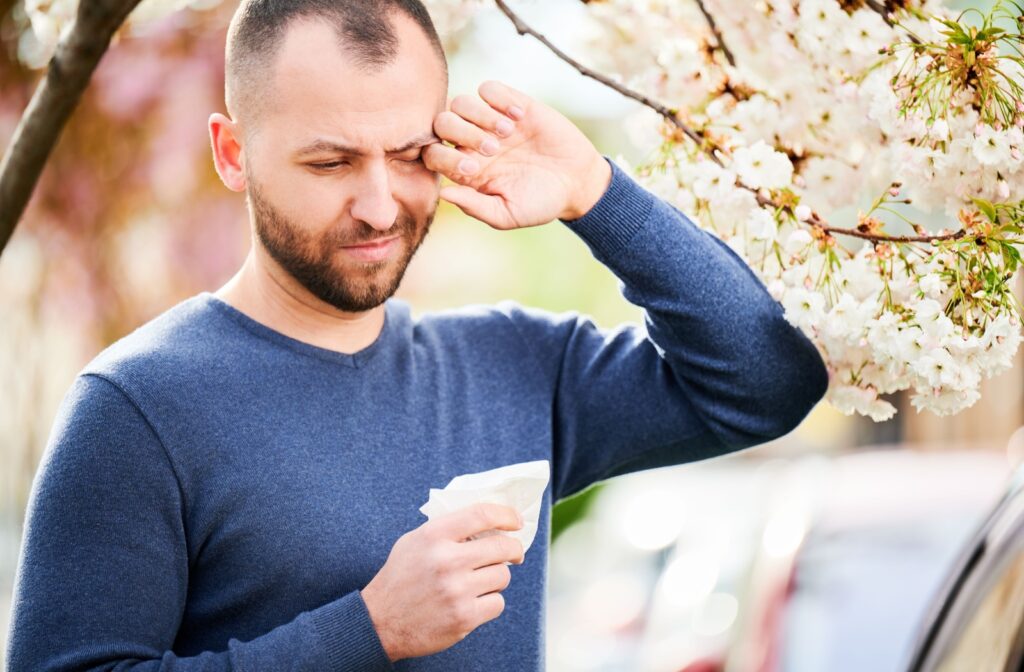 A young adult holding a tissue while standing under a tree outdoors, rubbing their eyes in discomfort due to dry eye and allergies.