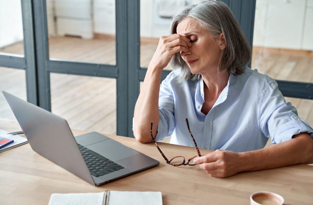 An elderly person with irritated eyes pinching the bridge of their nose at their desk with a laptop.