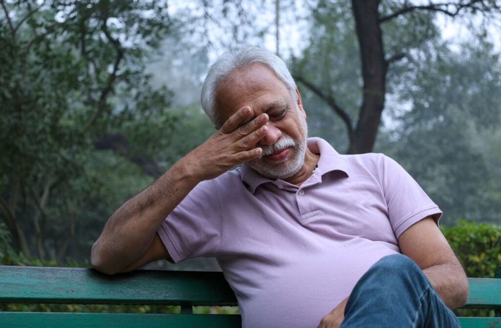 Older adult sitting on a bench outdoors in a foggy forest area, rubbing their eyes with one hand.