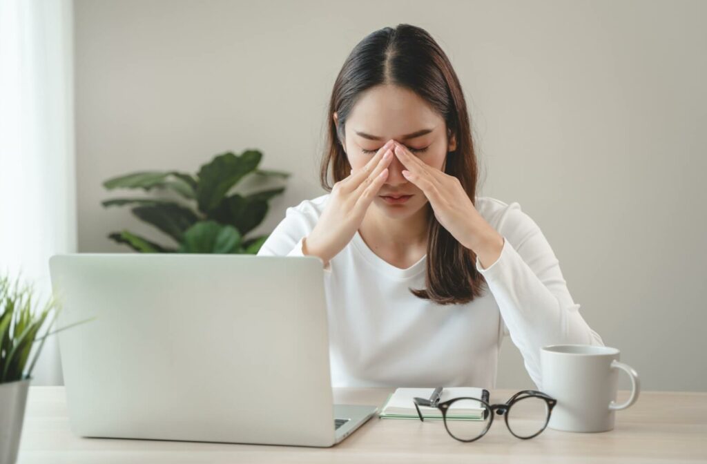 A person sitting at a desk with a laptop, pressing their fingers to the bridge of their nose as if experiencing eye strain or discomfort.