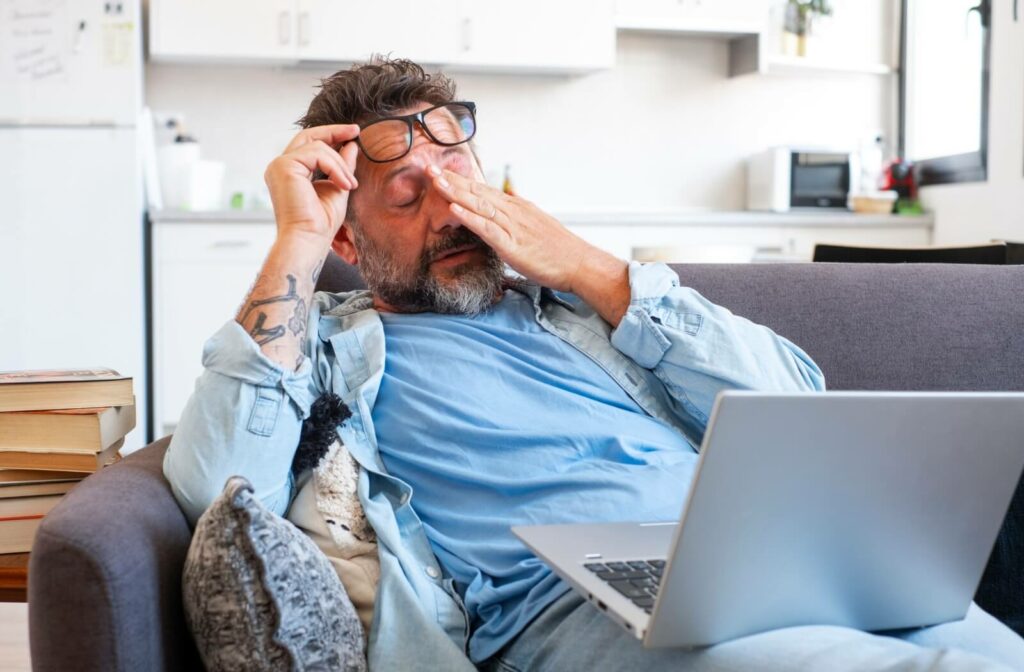 A person sitting on a couch with a laptop, lifting their glasses and rubbing their eyes as if feeling tired or experiencing eye discomfort.